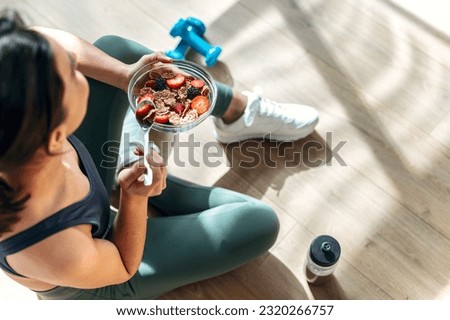 Shot of athletic woman eating a healthy bowl of muesli with fruit sitting on floor in the kitchen at home Royalty-Free Stock Photo #2320266757