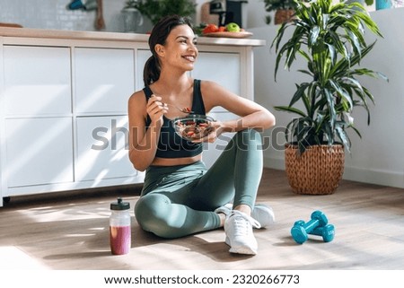 Shot of athletic woman eating a healthy bowl of muesli with fruit sitting on floor in the kitchen at home Royalty-Free Stock Photo #2320266773