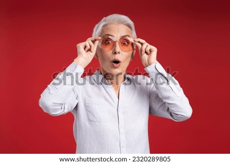 Excited gray haired woman wearing sunglasses, looking at camera isolated on red background
