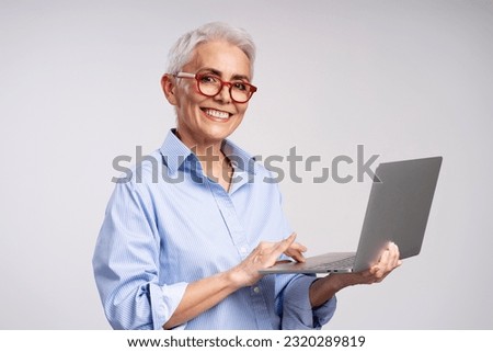 Portrait of smiling beautiful mature woman wearing stylish red glasses, using laptop, looking at camera isolated on gray background. Gray haired businesswoman, successful business concept