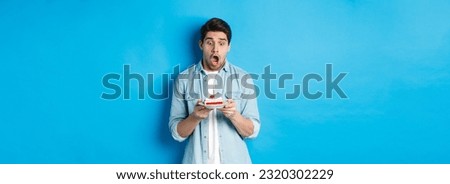 Man looking shocked at birthday cake, standing against blue background.