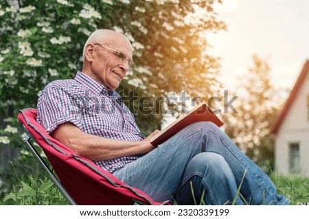 Retired man seated on armchair wearing glasses reading book in garden outdoor of cottage house. Royalty-Free Stock Photo #2320339199
