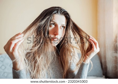 Young annoyed woman is upset about dirty oily and greasy hair touching it at home. Health treatment. Hair care products. Selfcare Royalty-Free Stock Photo #2320341003