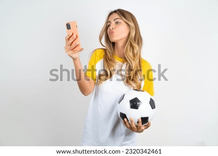 Young beautiful woman wearing football T-shirt blows air kiss at camera of smartphone and takes selfie, sends mwah via online call.