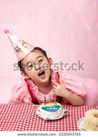A toddler from Asia (Korean-Indonesian) is wearing a party dress and a birthday hat, with an expressive face while looking at a cake in front of them. The portrait is isolated with a pink background.