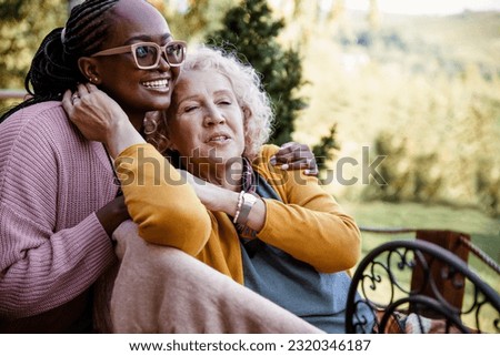 Mixed race female family relaxing at the patio of the country house. Young African American daughter with disabled mature Caucasian mother on sunny day outdoors on porch. Women friends care Royalty-Free Stock Photo #2320346187