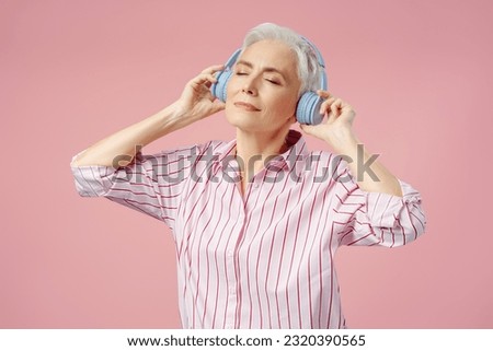 Smiling stylish senior woman with eyes closed listening music wearing wireless headphones, dancing, having fun isolated on pink background. Technology, positive lifestyle concept 