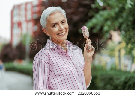 Cheerful stylish gray haired woman looking at camera while eating tasty ice cream in park. Positive lifestyle and tasty food concept 