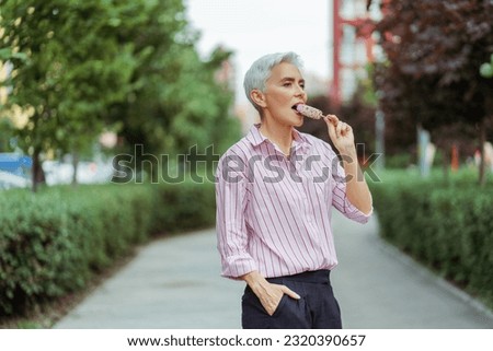 Relaxed happy gray haired woman eating tasty ice cream and looking away standing in park. Summer vacation, positive lifestyle concept 