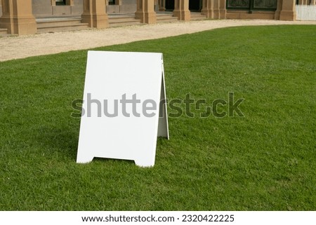 Background texture of a blank white advertising information board place outdoor on green and healthy grass lawn. Mockup template of a event welcome sign with an old mansion at the back.copy space