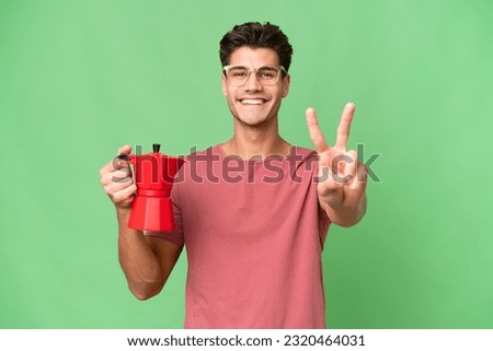 Young caucasian man holding coffee pot over isolated background smiling and showing victory sign