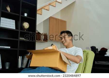 Smiling young man customer open parcel cardboard box sit on sofa at home,  consumer unpacking carton package looking inside receive good purchase post mail shipping delivery concept.