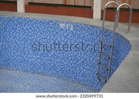 Swimming pool iron stair reach down to the pool. Semicircular metal steps of ladder.  blue colour mosaic.  large dry, dirty  pool without water. 