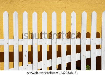 white painted wooden picket fence with shadows and a yellow painted wall Royalty-Free Stock Photo #2320538431