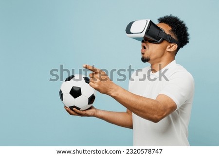 Young shocked man fan wearing basic t-shirt cheer up support football sport team hold in hand soccer ball watching in vr headset pc gadget watch tv live stream isolated on plain blue color background