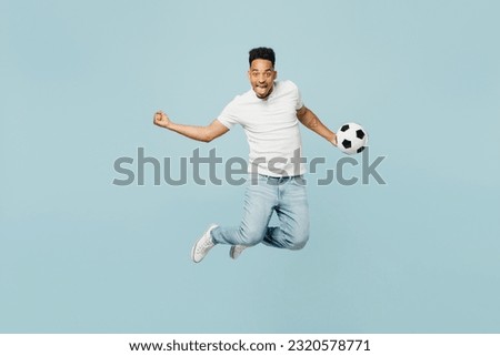 Full body overjoyed young man fan wears t-shirt do winner gesture cheer up support football sport team hold soccer ball jump high watch tv live stream isolated on plain pastel blue color background
