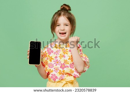 Little child kid girl 6-7 years old wears casual clothes hold use blank screen workspace area mobile cell phone do winner gesture isolated on plain green background. Mother's Day love family concept
