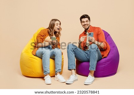 Full body amazed young couple two friends family man woman wear casual clothes together sit in bag chair hold in hand use mobile cell phone isolated on pastel plain light beige color background studio