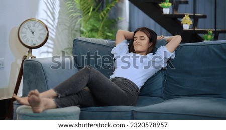 Happy young beautiful smiling woman sitting resting  on sofa raising arms stretching back muscles enjoying peaceful weekend at home. Indian female leaning on couch with closed eyes  hands behind head Royalty-Free Stock Photo #2320578957