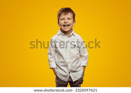 Cute little boy in trendy white shirt smiling broadly while keeping hands in pockets against yellow background Royalty-Free Stock Photo #2320597621