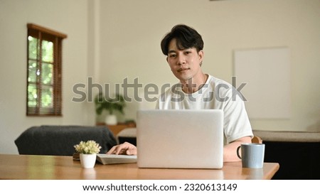 A handsome young Asian man in casual clothes sits at a table with his laptop in his minimalist living room.