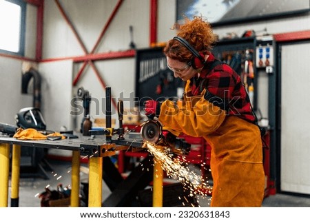 Woman in STEM using grinder to cut a metal bar, wearing gloves and a apron with protective sleeves. She's making a new metal part for her engineering project