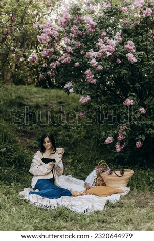 young beautiful brunette woman sits on a picnic blanket near a lilac in the park. woman drinking coffee or tea in cermic cup outdoors. picnic breakfast