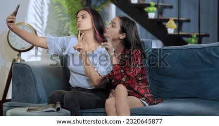 Smiling female sitting on sofa take funny faces selfie with female girlfriend use mobile phone at indoor home. Happy Indian girls making self photos on smartphone blowing kiss enjoying together 