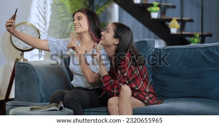Smiling female sitting on cosy sofa take funny faces selfie with female girlfriend use mobile phone at indoor home. Happy Indian girls making self photos on smartphone enjoy together 