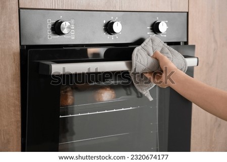 Woman opening electric oven with buns in kitchen, closeup Royalty-Free Stock Photo #2320674177