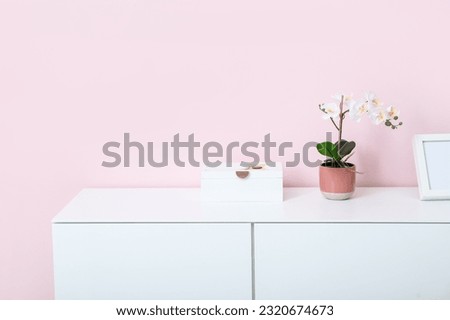 White chest of drawers with orchid flower and blank picture near pink wall in room