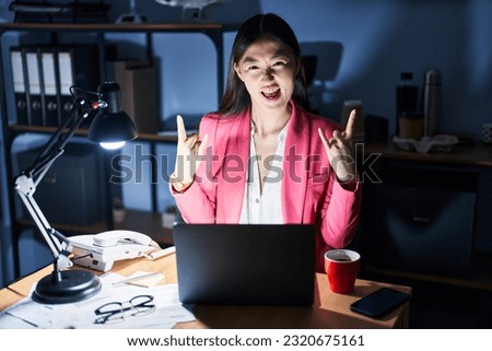Chinese young woman working at the office at night shouting with crazy expression doing rock symbol with hands up. music star. heavy concept. 
