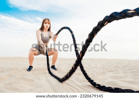 Muscular young concentrated woman working out with battle ropes on the beach, intense functional circuit training. Doing sport outdoors. Crossfit, fitness and workout concept. Selective focus Royalty-Free Stock Photo #2320679145