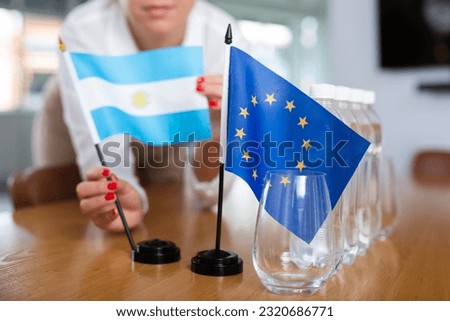 Young woman in business clothes puts flags of Argentina and European Union on negotiating table in office