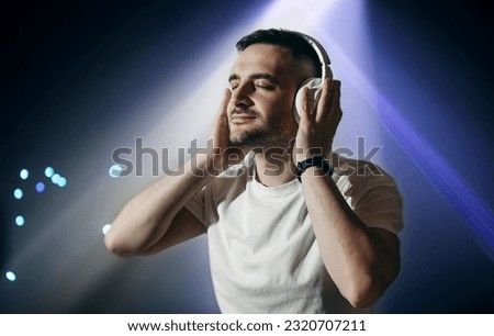 Young bearded man with headphones listens to music on dark blue background in nightclub with lights.