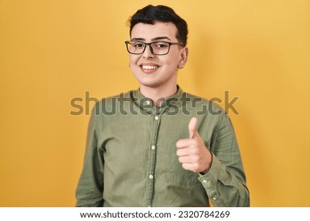 Non binary person standing over yellow background doing happy thumbs up gesture with hand. approving expression looking at the camera showing success. 