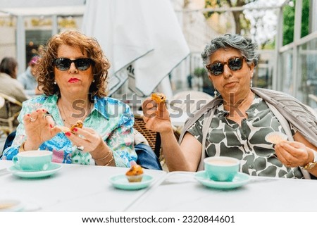 Positive mature female friends in trendy outfit and sunglasses sitting at table with cup of coffee and eating snacks while looking at camera outdoors