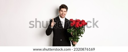 Image of handsome romantic guy in black suit, holding bouquet of roses and pointing at camera, congratulating with holiday, standing against white background.