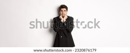 Image of scared and insecure young businessman in suit, trembling from fear and looking horrified, standing over white background.