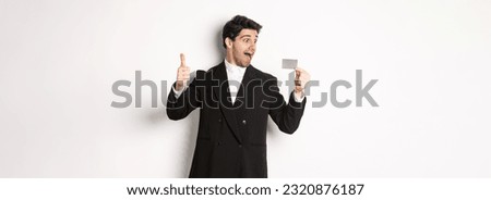 Image of excited handsome businessman, showing credit card and thumb-up, standing against white background.