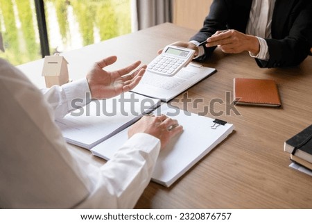 Female real estate agent in suit showing numbers and calculating house, property price on calculator for buyer or client while sitting at desk to discuss and negotiate about contract at office. Royalty-Free Stock Photo #2320876757
