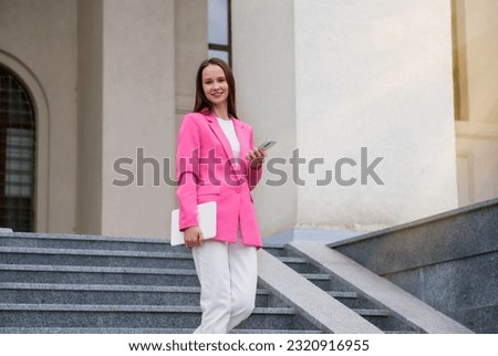 Young happy elegant stylish professional business woman, business lady executive manager in pink suit holding smartphone standing outdoor using mobile corporate app on mobile phone.