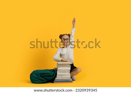 Cute Little Nerdy Schoolgirl Sitting With Pile Of Books And Raising Hand, Smiling Preteen Female Child Answering Question On Lesson, Enjoying Knowledge And Study, Posing On Yellow Background