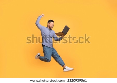 Emotional young asian man jumping up with laptop in hand, joyful millennial man looking at computer screen and shaking fist, celebrating online win, having fun on yellow studio background, copy space