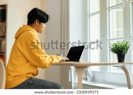 Homeschooling concept. Japanese teen student boy studying online, typing on laptop sitting indoors. Guy using portable pc to make homework at desk, learning from home. Side view Royalty-Free Stock Photo #2320939571