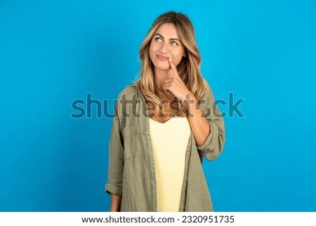 Lovely dreamy young beautiful blonde woman wearing overshirt keeps finger near lips looks aside copy space.