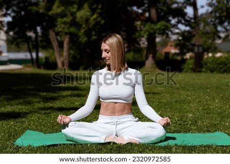 young beautiful blond woman in white sportswear practice yoga on green mat in lawn outdoors in city park in summer, sitting in lotus position, green trees in background, relaxation concept