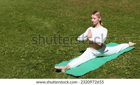 young beautiful blond woman in white sportswear practice yoga on green mat in lawn outdoors in city park in summer, sitting in longitudinal twine, relaxation concept, copy space
