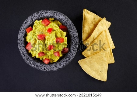 Top view of a delicious avocado guacamole with red diced tomatoes in a typical stone Mexican mortar or molcajete by a few totopos or nachos over a black background. Royalty-Free Stock Photo #2321015149