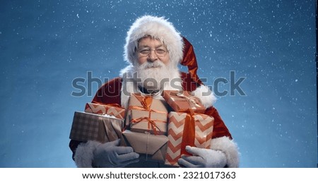 Close up shot of senior bearded man in santa clause outfit carrying gift boxes, isolated on blue background - christmas spirit, holiday mood